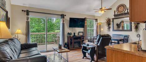 A spacious living room with a black leather sofa, a wooden coffee table, and a fireplace. There is a television mounted on the wall, a dining area with a table and chairs, and a large window with curtains.