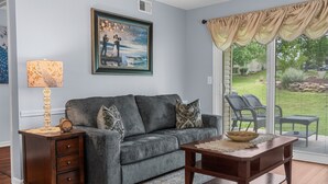 A living room with a gray sofa, a wooden side table, a lamp, a framed painting, and a view of the outdoors.