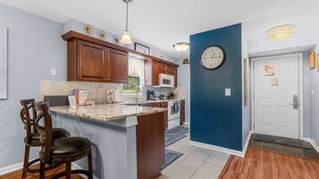A kitchen with wooden cabinets, a marble countertop, a built-in microwave, and a clock on the wall.
