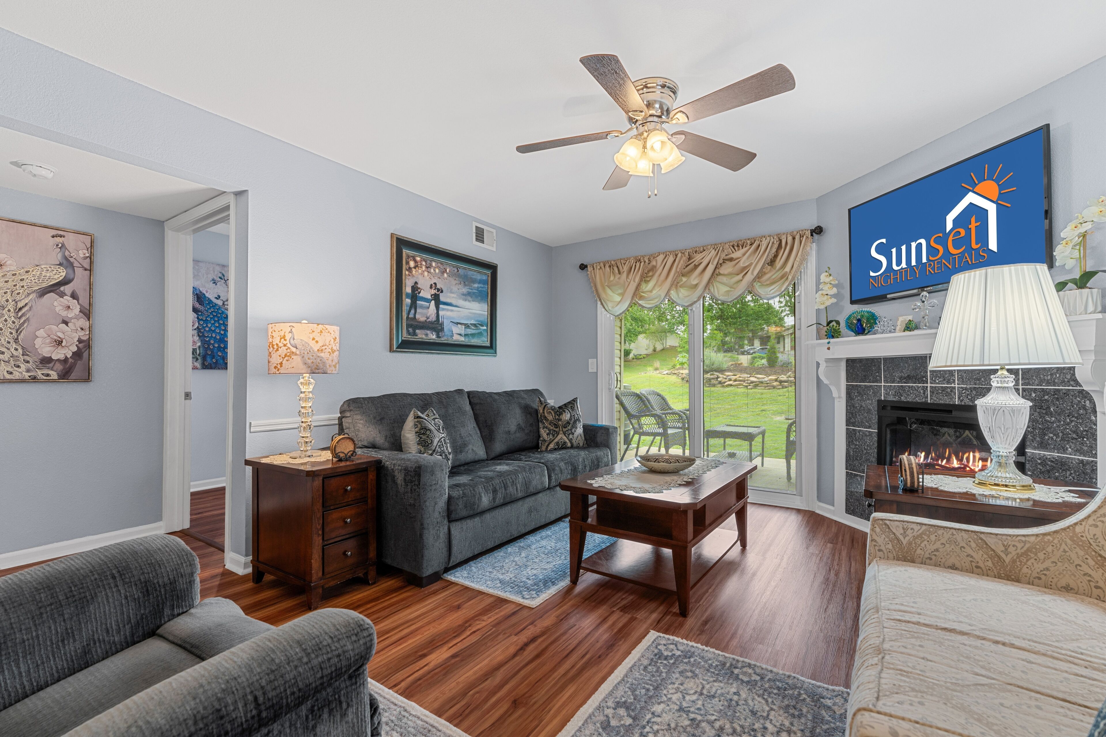 A living room with a sofa, coffee table, and fireplace, a ceiling fan, and a TV displaying 'Sunset Nightly Rentals'.
