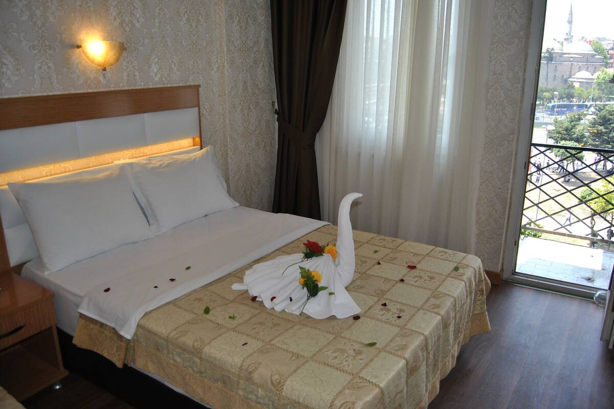 A hotel room with a bed, a nightstand, a window with curtains, and a view of a building outside.