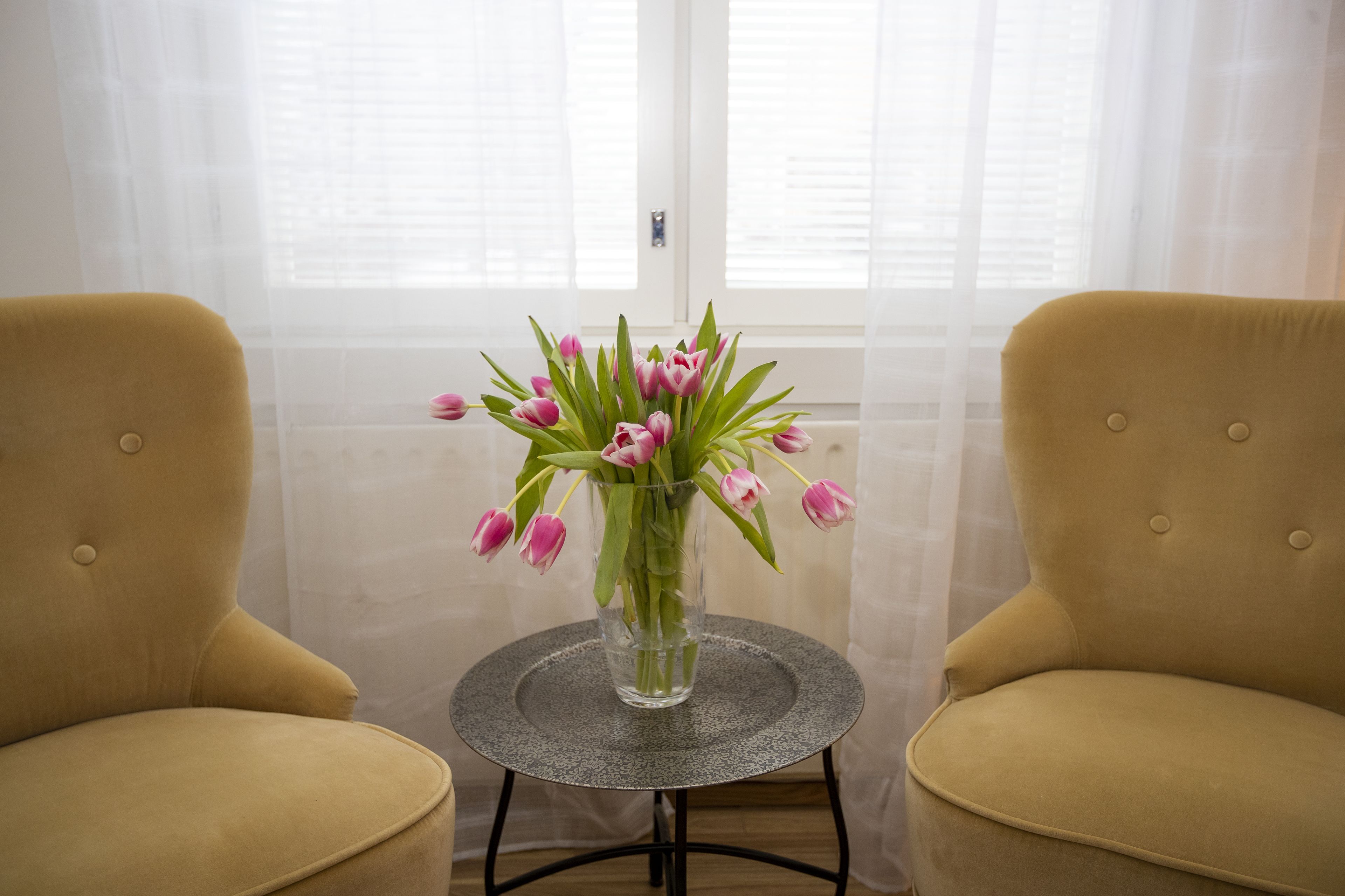 Een kamer met twee gele stoelen, een klein rond tafeltje met een vaas roze tulpen en een raam met dunne gordijnen.