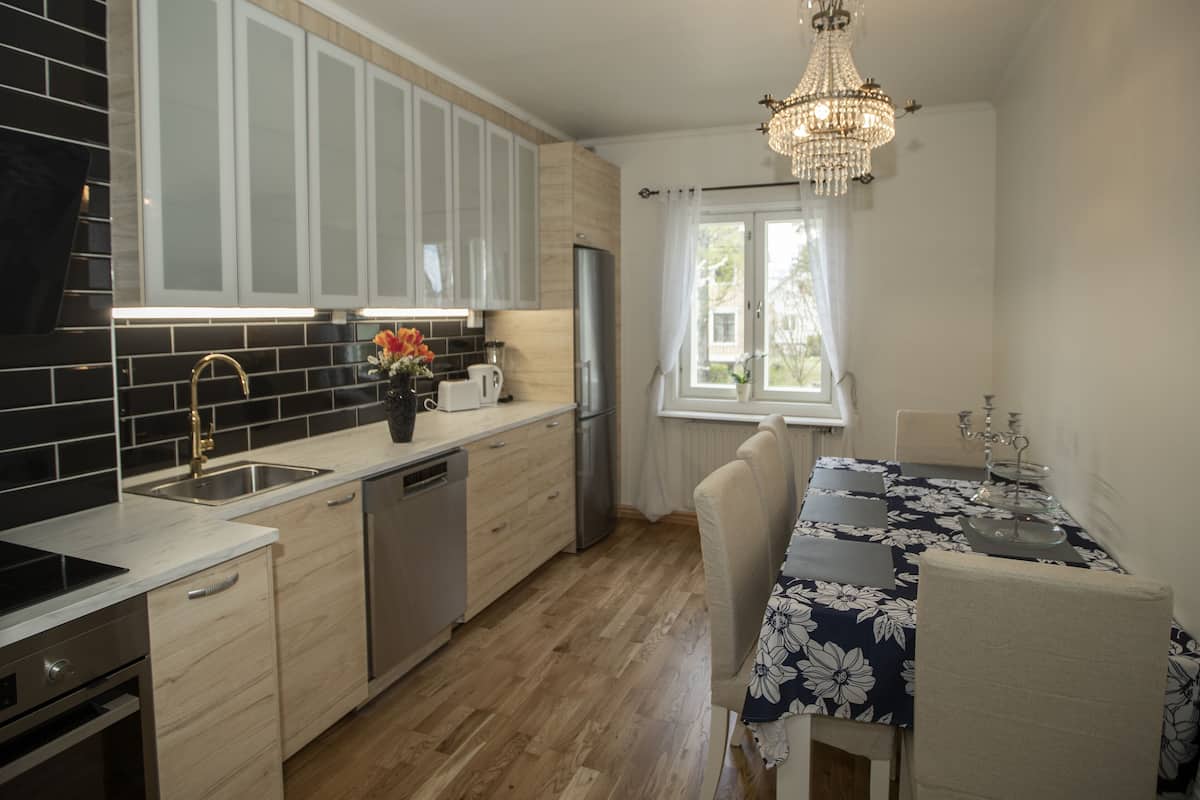 A modern kitchen with a built-in oven, a dishwasher, and a stainless steel refrigerator. There is a dining table with a floral tablecloth and chairs. A chandelier hangs from the ceiling, and a window with curtains is visible.
