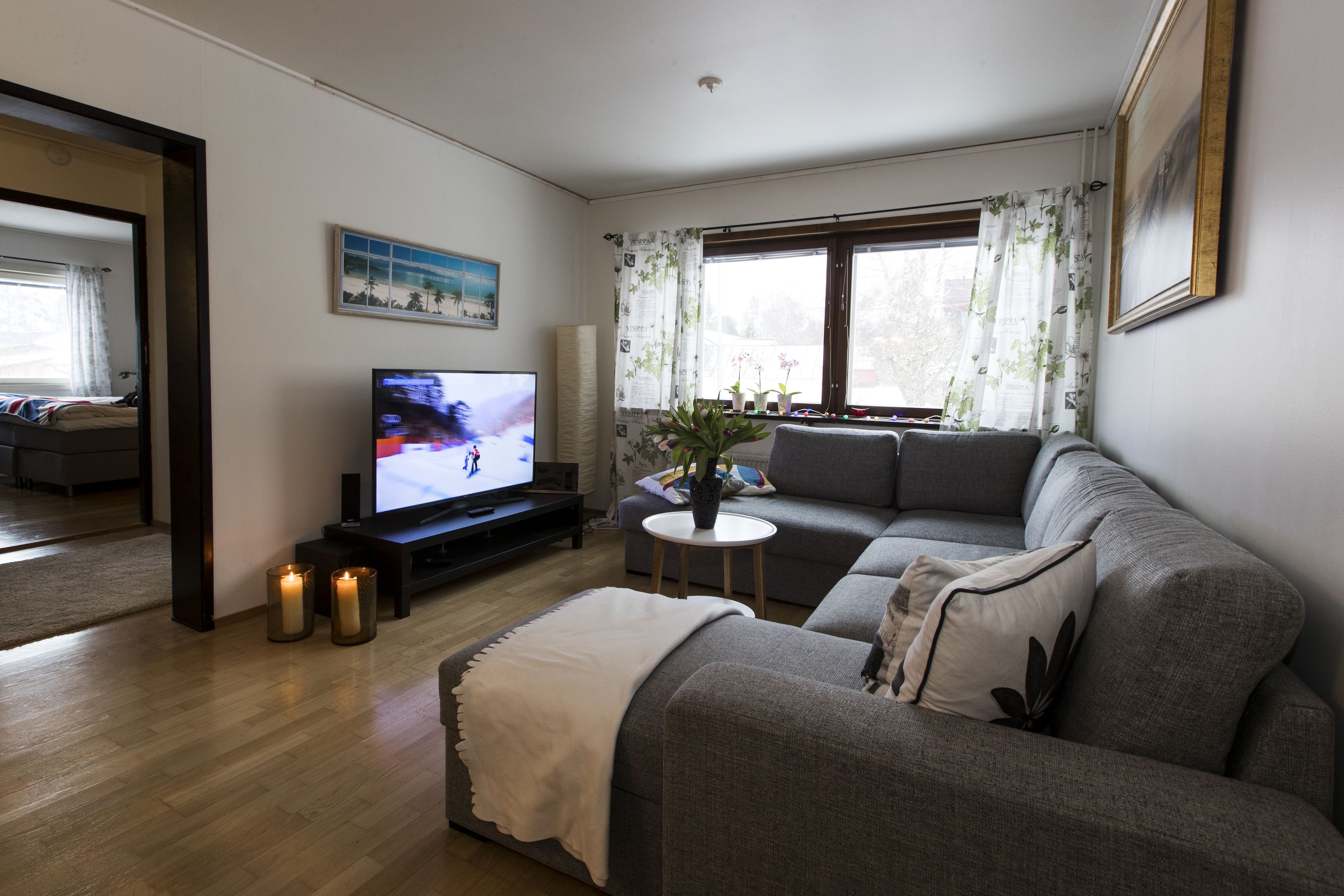 Una sala de estar con un sofá gris, una mesita redonda pequeña, un televisor y una ventana con cortinas florales.