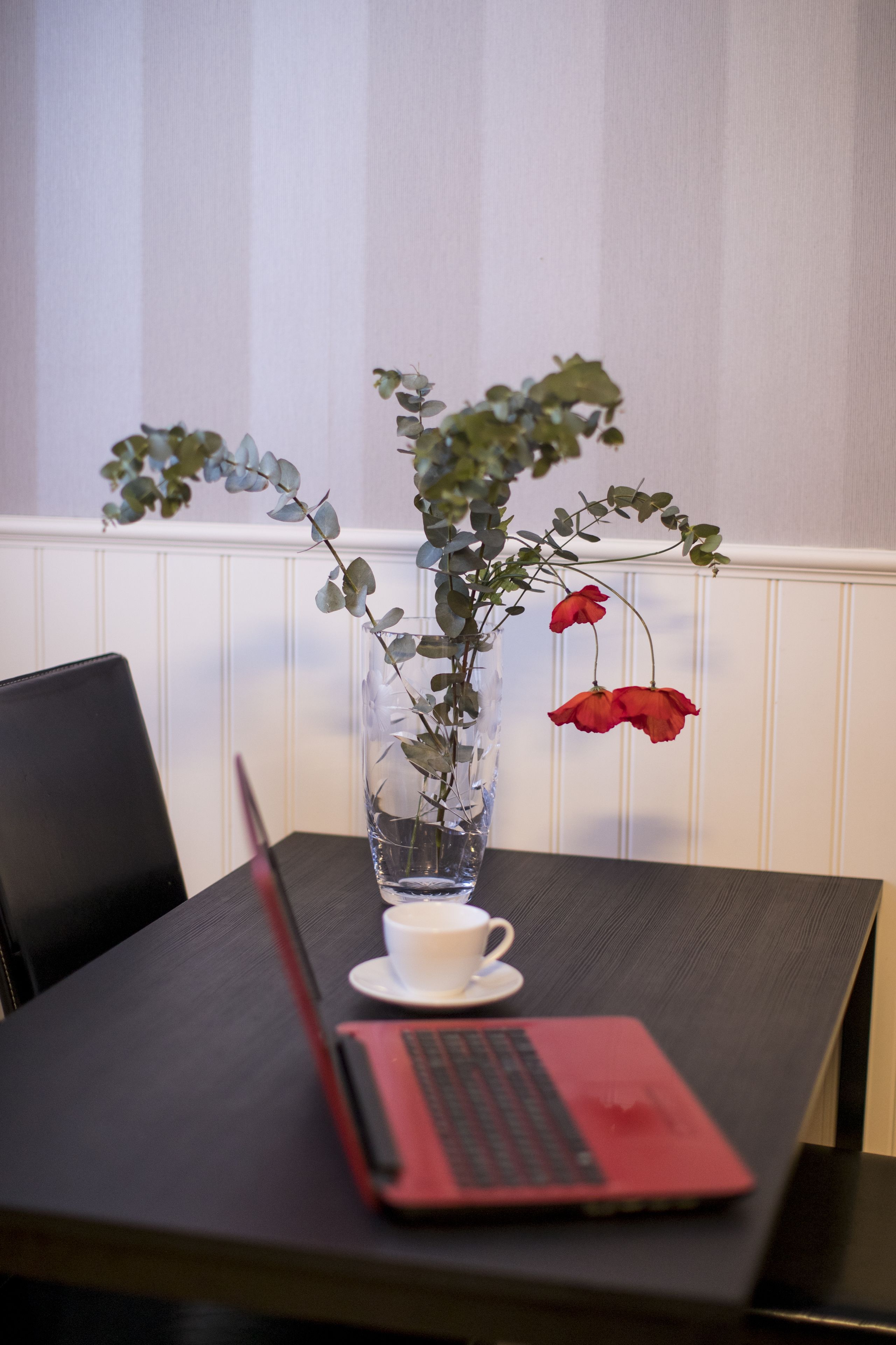 A laptop on a table with a white cup and a vase with green leaves and red flowers.