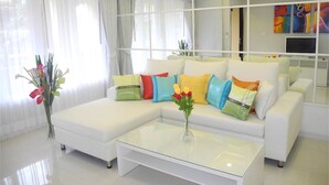 A modern living room with a white sectional sofa, colorful cushions, a glass coffee table, and a large mirror reflecting the room. - Kata Plaza 1 bedroom Centrical Apartment (Karon)
