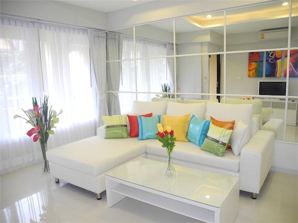A modern living room with a white sectional sofa, colorful cushions, a glass coffee table, and a large mirror reflecting the room. - Kata Plaza 1 bedroom Centrical Apartment (Karon)