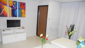 A modern living room with a flat-screen TV, white sofa, and a coffee table with a vase of flowers. - Kata Plaza 1 bedroom Centrical Apartment (Karon)