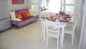 A compact living space with a purple sofa, white dining table, and chairs. A refrigerator is visible on the left. - Kata Plaza 1 bedroom Centrical Apartment (Karon)