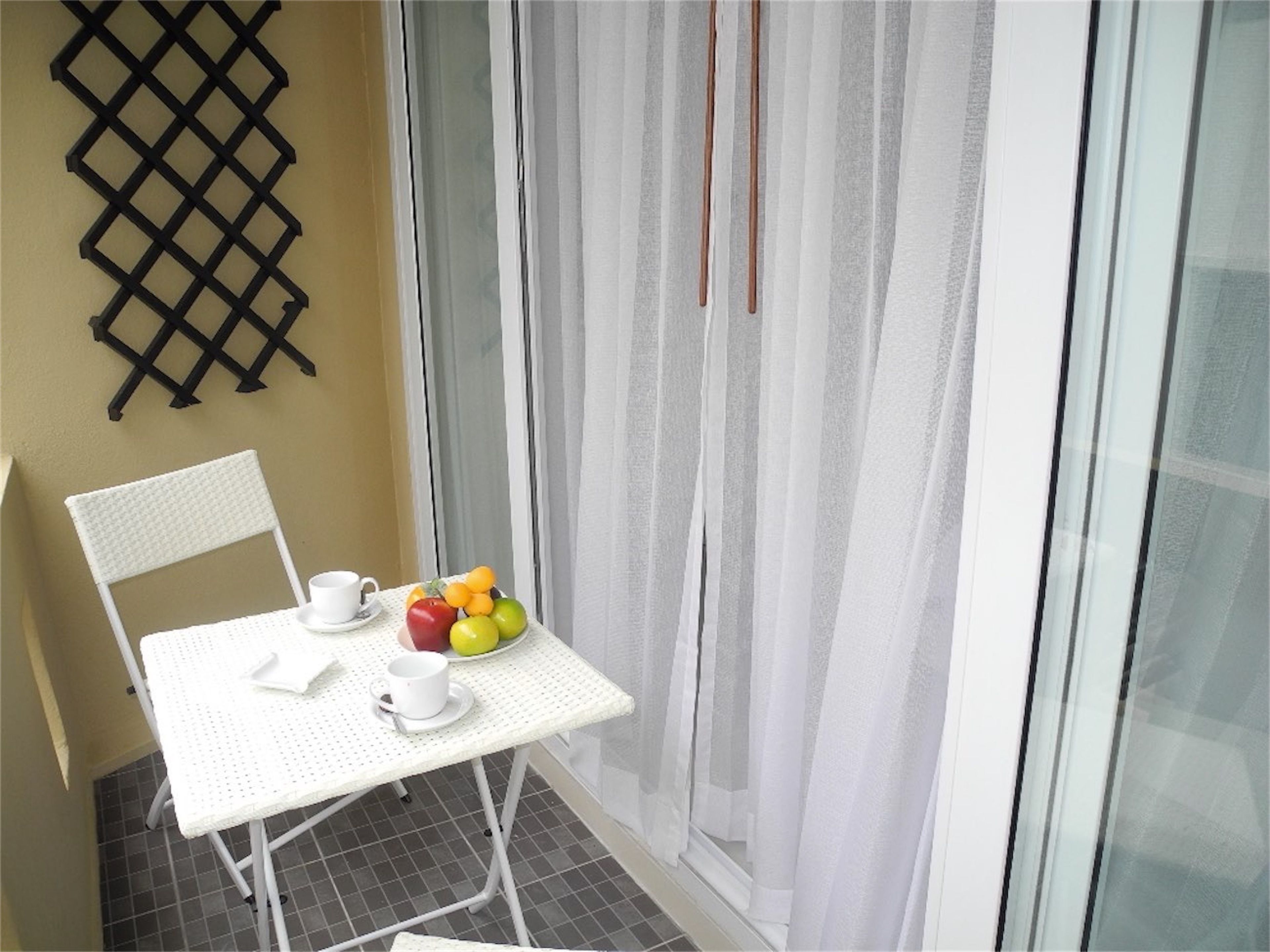 Un petit balcon avec une table blanche, deux chaises, un panier de fruits et une tasse.