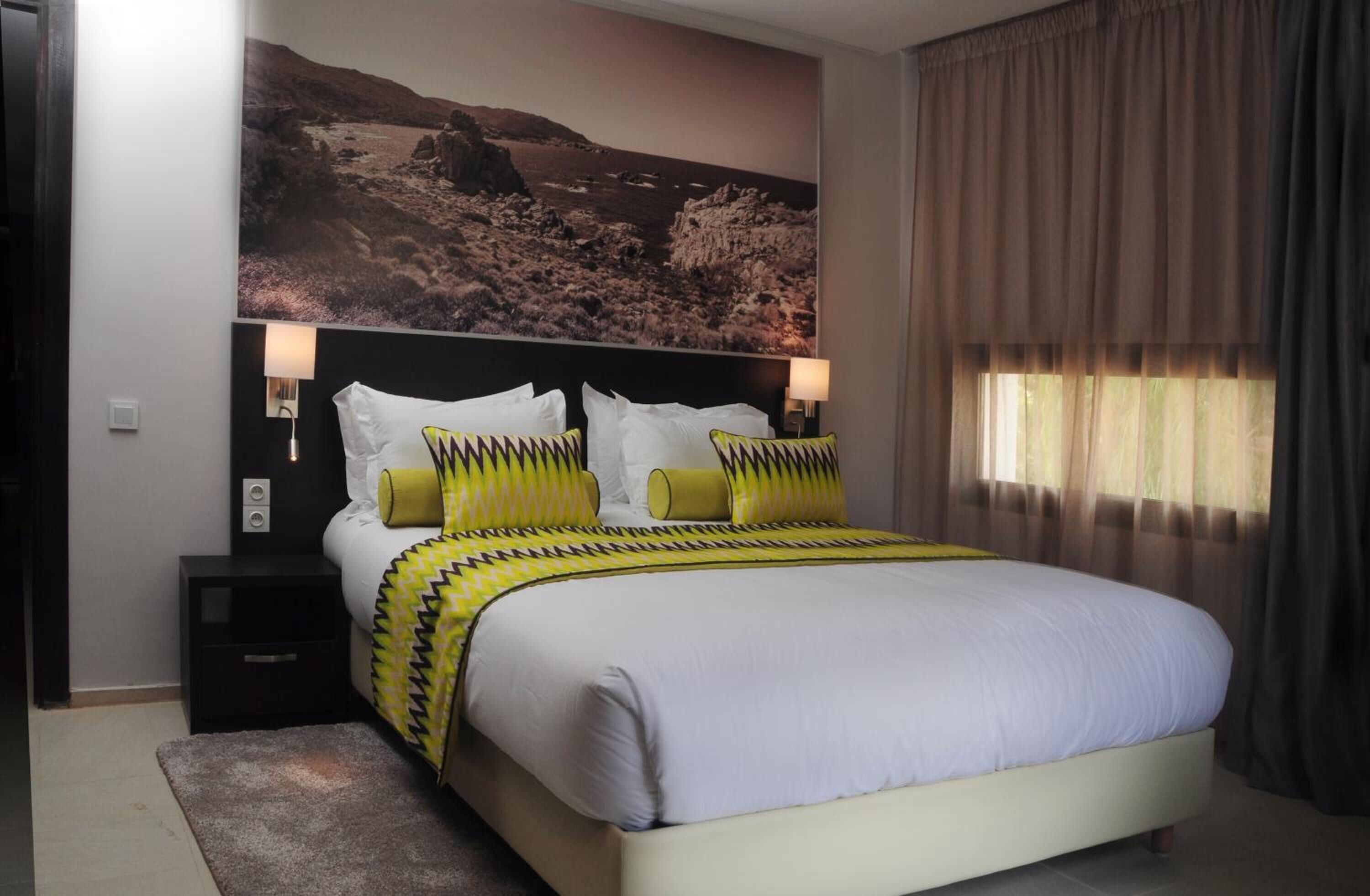 Un dormitorio con cama, mesitas de noche, un cuadro de paisaje grande y una ventana con cortinas.