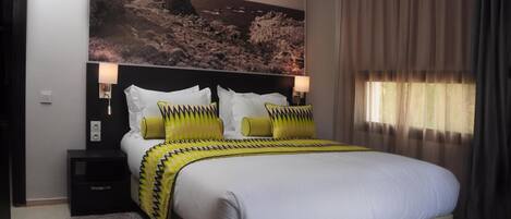 Un dormitorio con cama, mesitas de noche, un cuadro de paisaje grande y una ventana con cortinas.