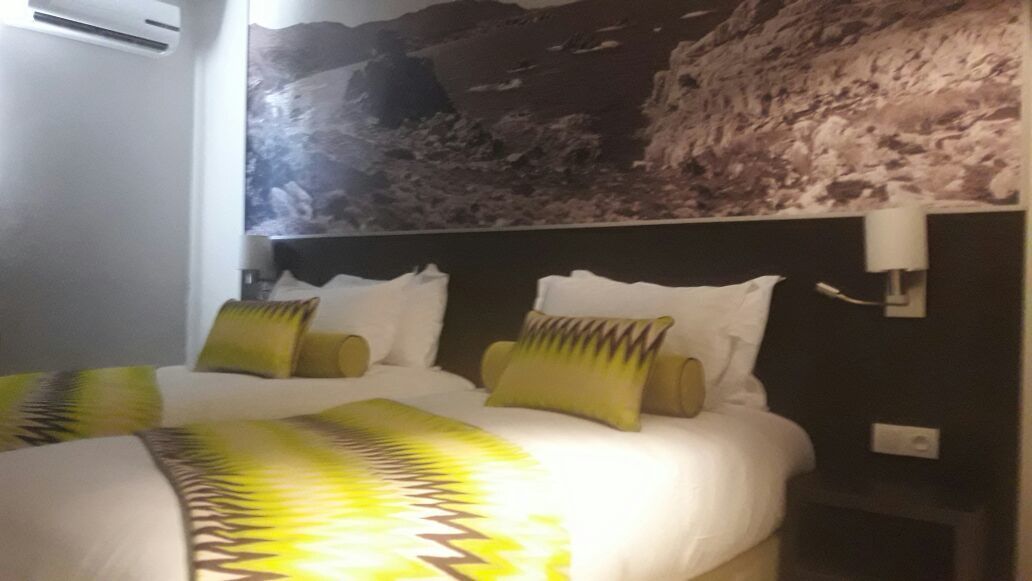 A hotel room with a bed, pillows, a wall-mounted air conditioner, and a painting of a rocky landscape.