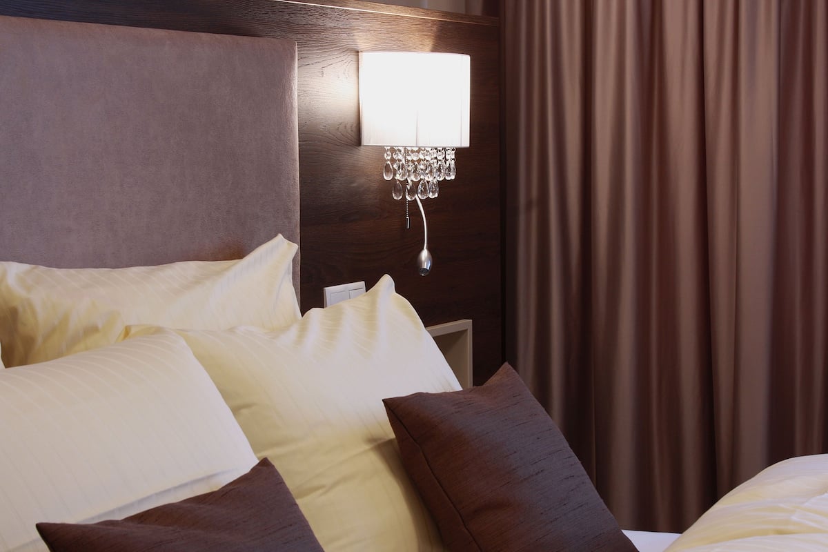 A hotel room with a neatly made bed, a wall-mounted lamp, and a curtain.