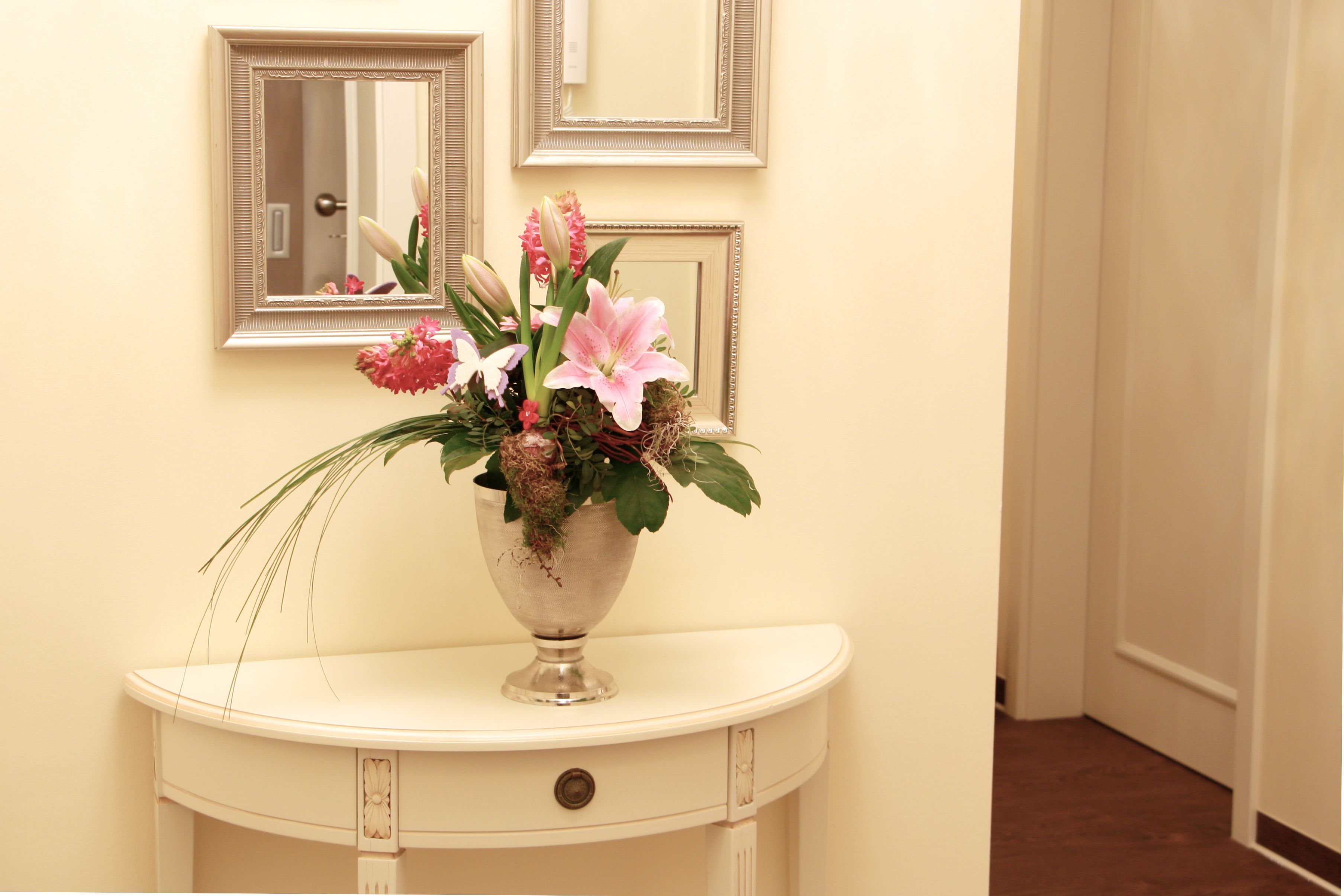Una consola blanca con un florero plateado que contiene flores rosas y blancas, frente a una pared color crema con dos espejos enmarcados.