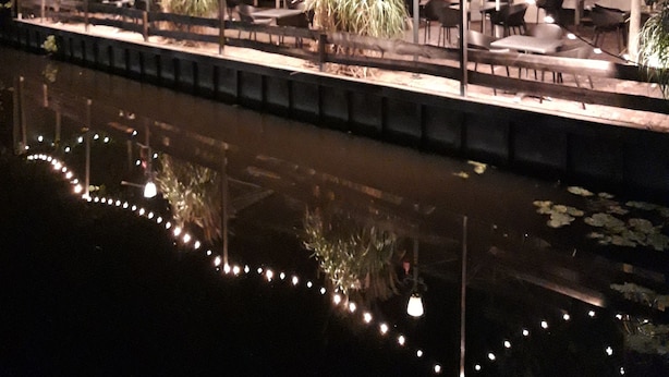 An outdoor restaurant with tables and chairs, string lights, and a water body in the foreground. - Abbey Abcoude (Abcoude)