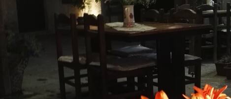 A dimly lit outdoor dining area with a table and chairs, surrounded by potted plants and flowers.