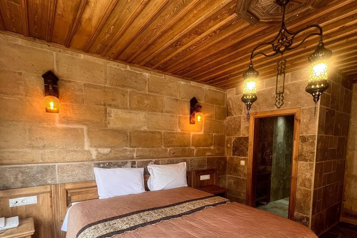 A bedroom with a wooden ceiling, stone walls, a bed with a pink bedspread, two bedside tables, and wall-mounted lamps.