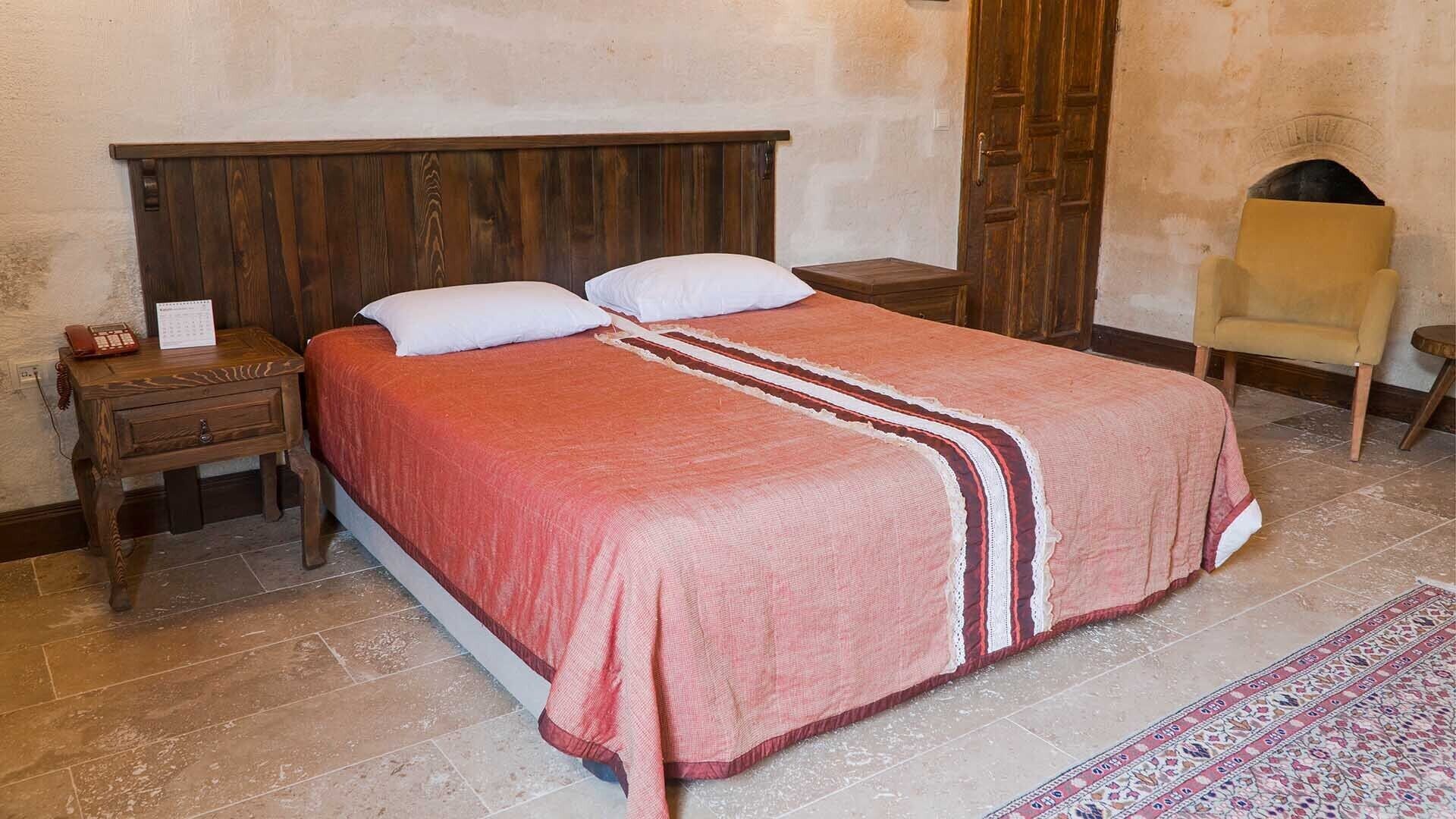 Ein Schlafzimmer mit einem Holz-Kopfteil, einem Bett mit einer rot-weiß gestreiften Tagesdecke, einem Nachttisch mit Telefon und Kalender, einem Sessel und einem gemusterten Teppich.
