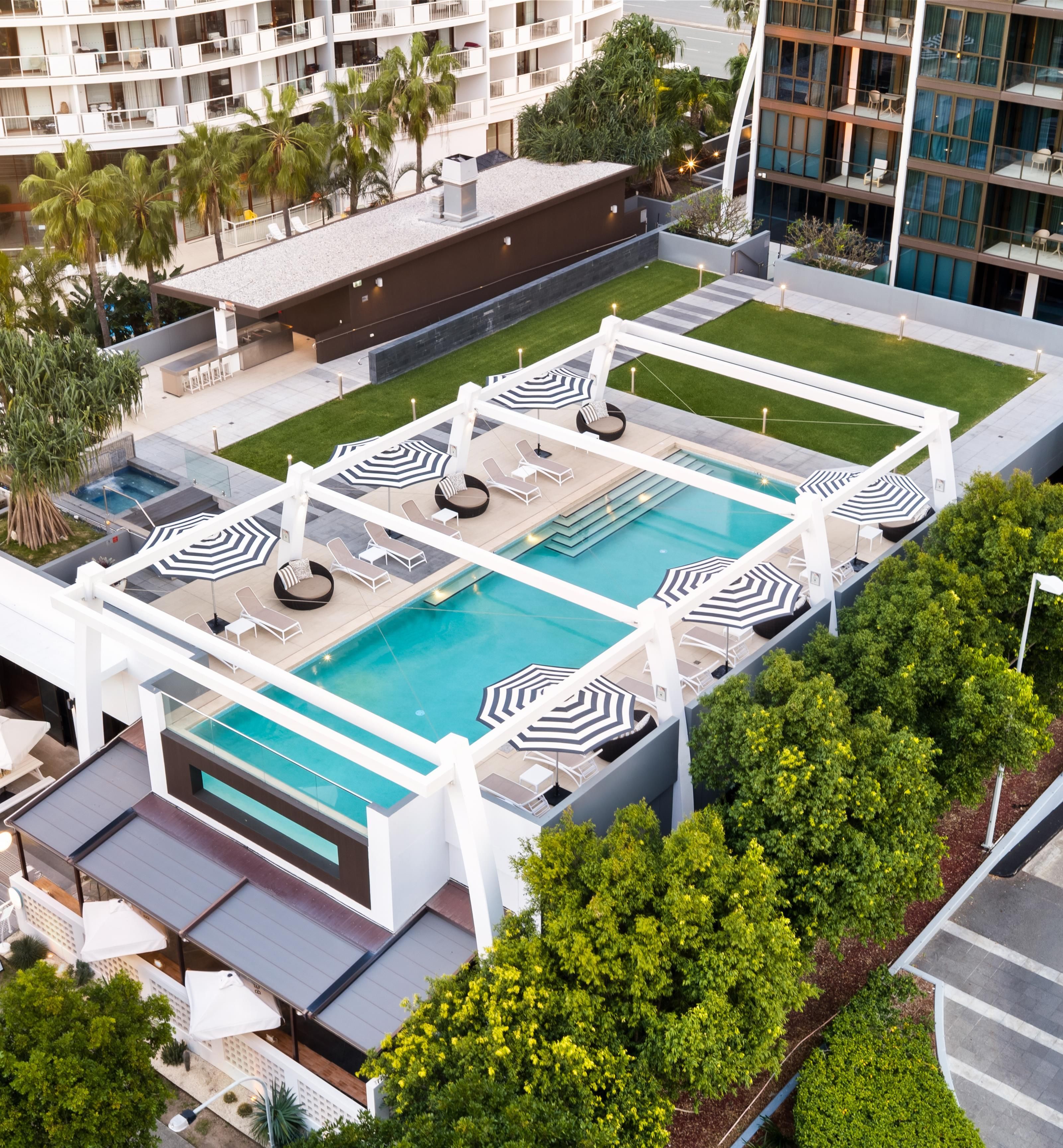 A rooftop pool area with multiple lounge chairs and umbrellas, surrounded by greenery and modern architecture.