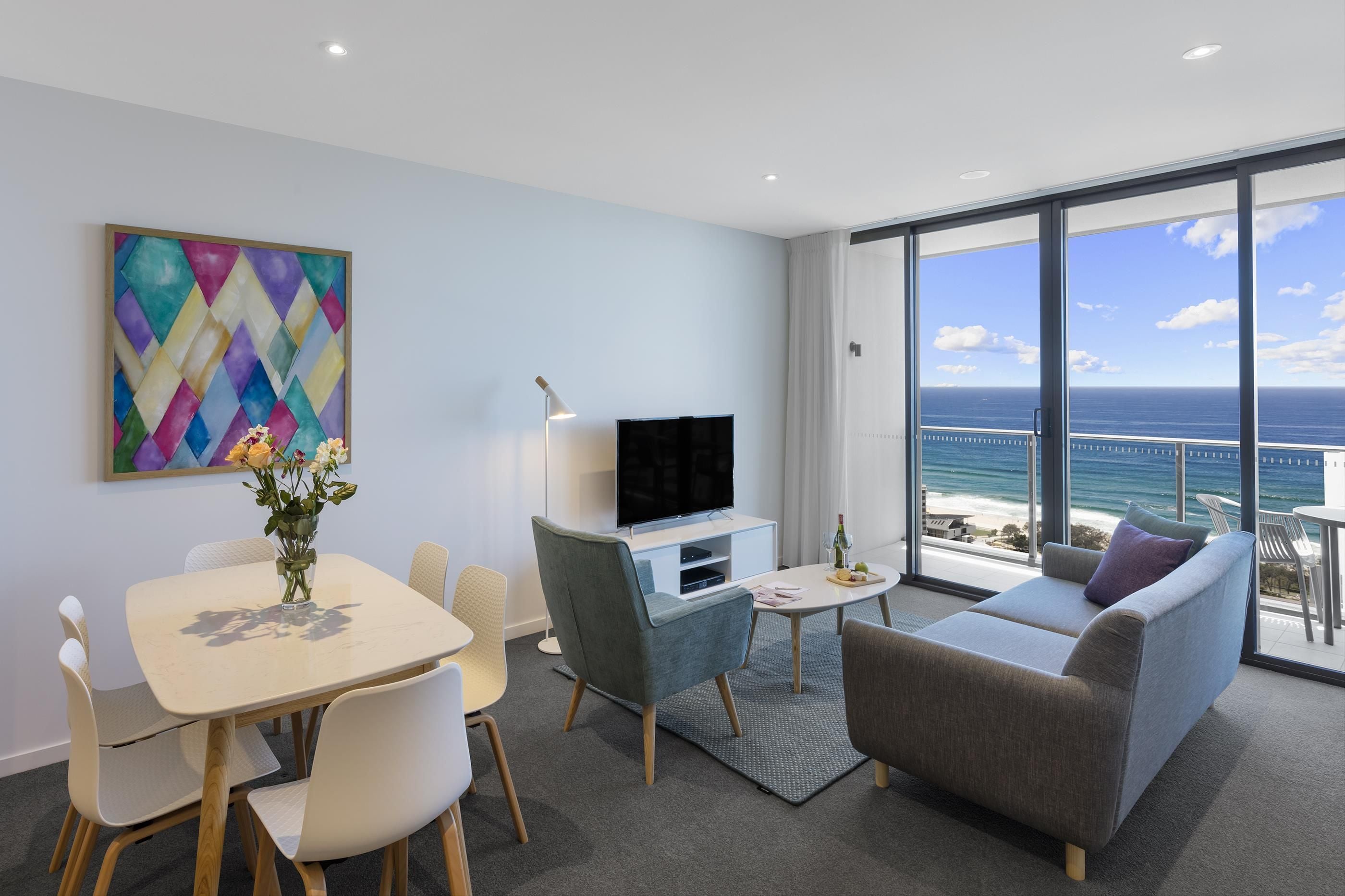 A modern living room with a flat-screen TV, a dining table with chairs, a sofa, and a large window offering a view of the ocean.