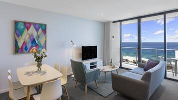 A modern living room with a flat-screen TV, a dining table with chairs, a sofa, and a large window offering a view of the ocean.