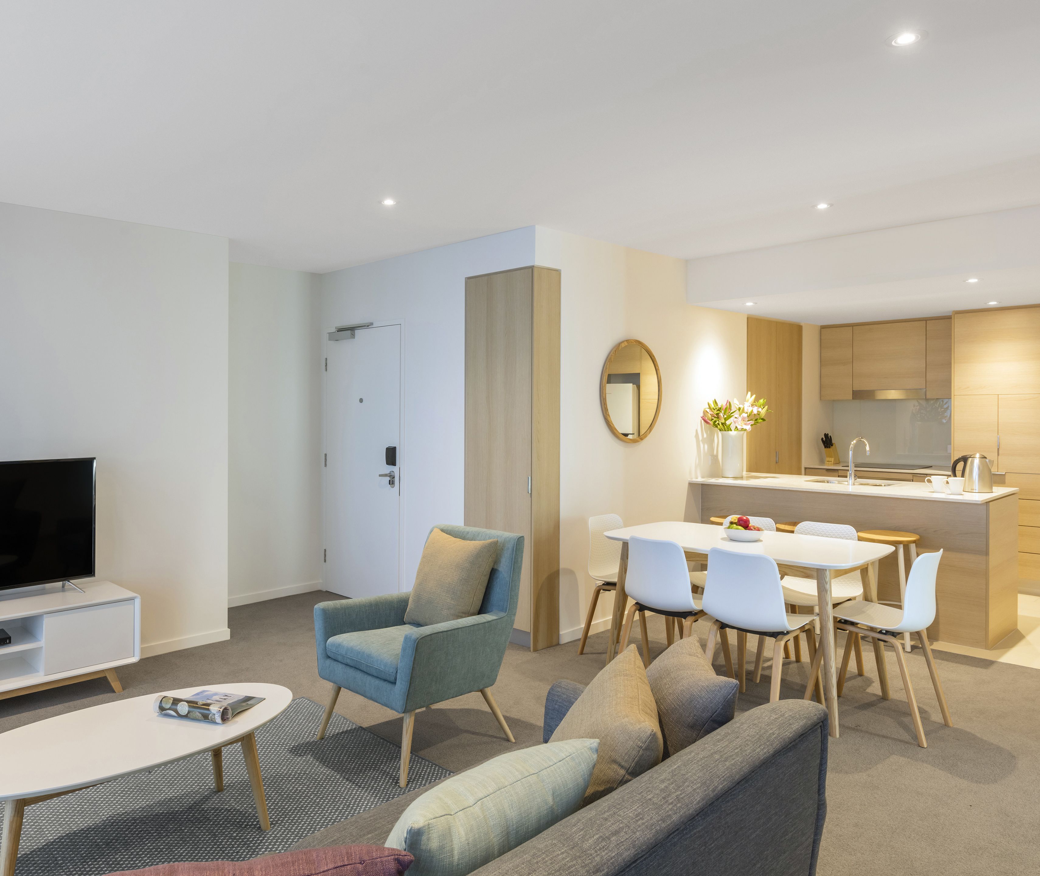 Un salon moderne avec un canapé, un fauteuil et un téléviseur, un coin repas avec des chaises et une table, et une cuisine avec des meubles en bois et un évier.