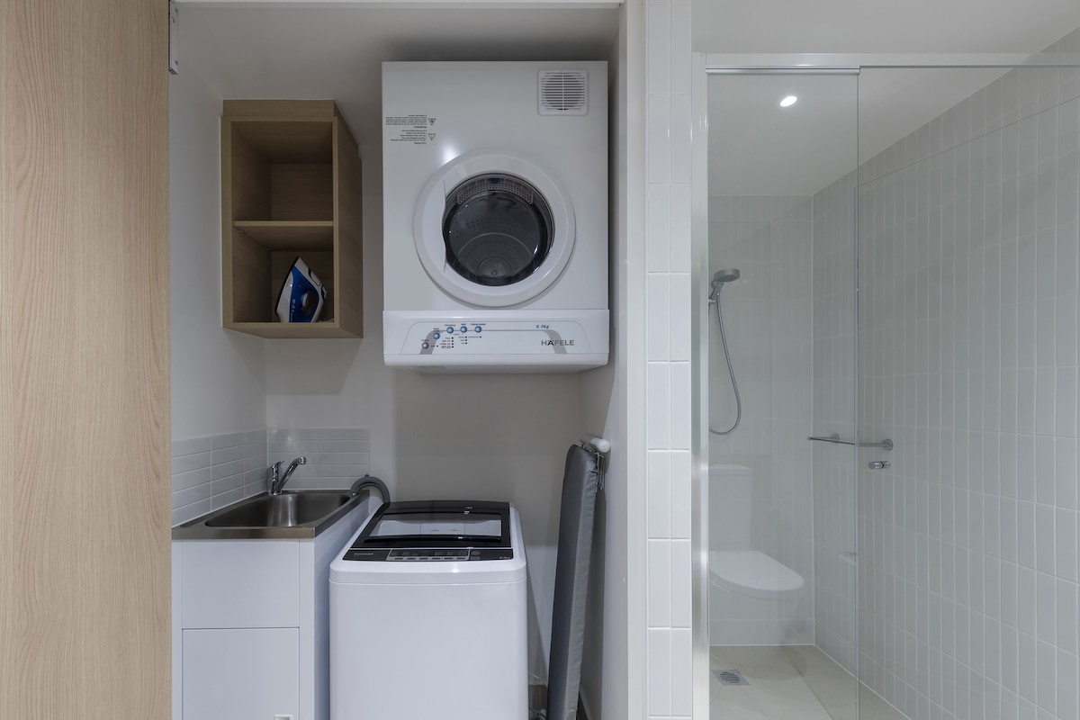 A compact laundry room with a front-loading washer and dryer, a small sink, and a glass-enclosed shower.