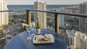 A table with a bottle of wine, glasses, and a cheese board with crackers, overlooking a cityscape and the ocean. - Avani Broadbeach Residences (Broadbeach)