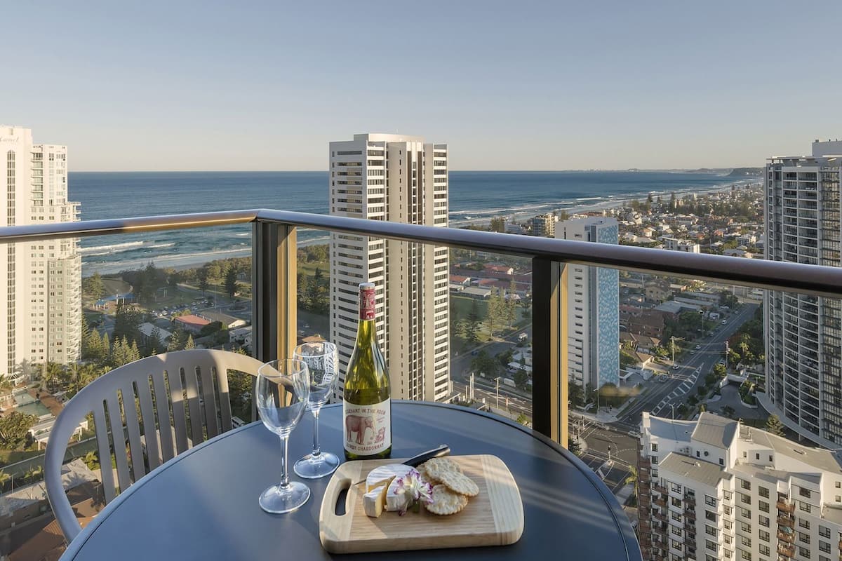 A table with a bottle of wine, glasses, and a cheese board on a balcony overlooking a cityscape and ocean.