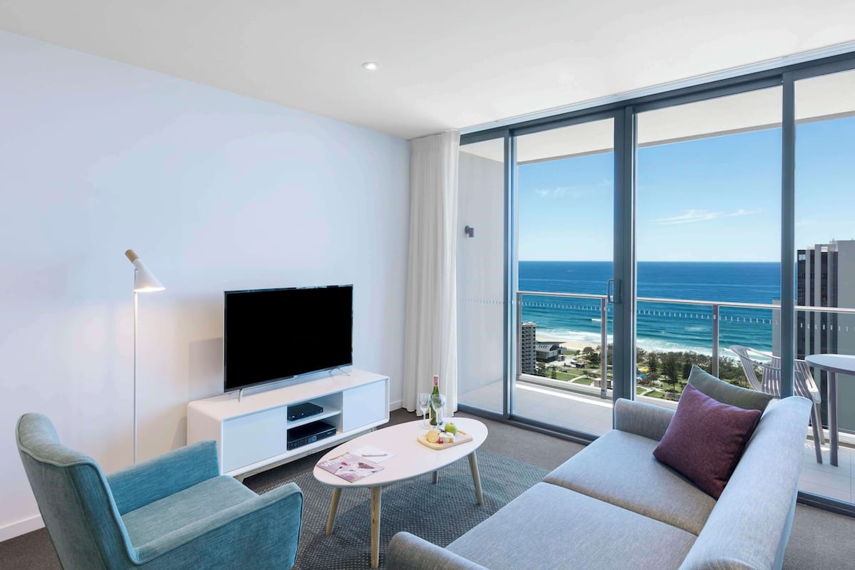 A modern hotel room with a large balcony, a flat-screen TV, a sofa, and a dining table.