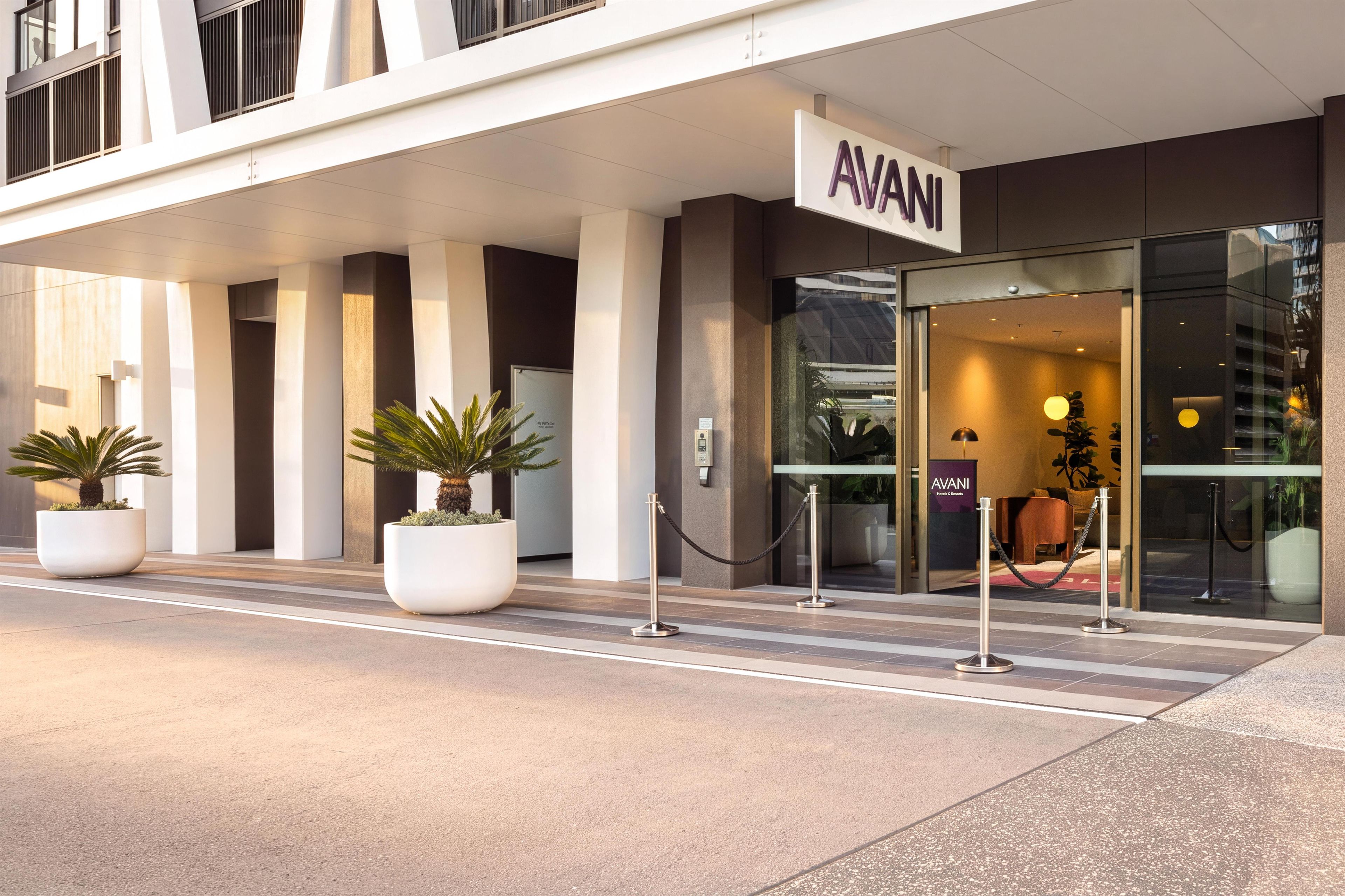A hotel entrance with a sign reading 'AVANI' and a glass door. There are potted plants and a security rope barrier in front.
