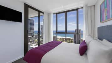 Una habitación de hotel con cama, televisor, un ventanal grande con vistas a la ciudad y al océano, y un cuadro en la pared.