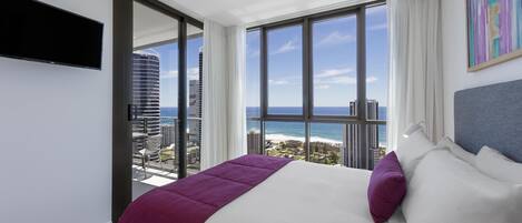 A hotel room with a bed, a TV, a large window with a city and ocean view, and a painting on the wall.