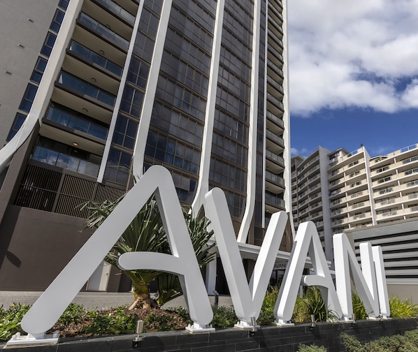 Un edificio moderno con letras blancas grandes que dicen "AVANI" en primer plano, rodeado de follaje y un cielo despejado.