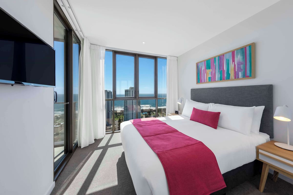 A hotel room with a large bed, a TV, a painting, and a view of the city.
