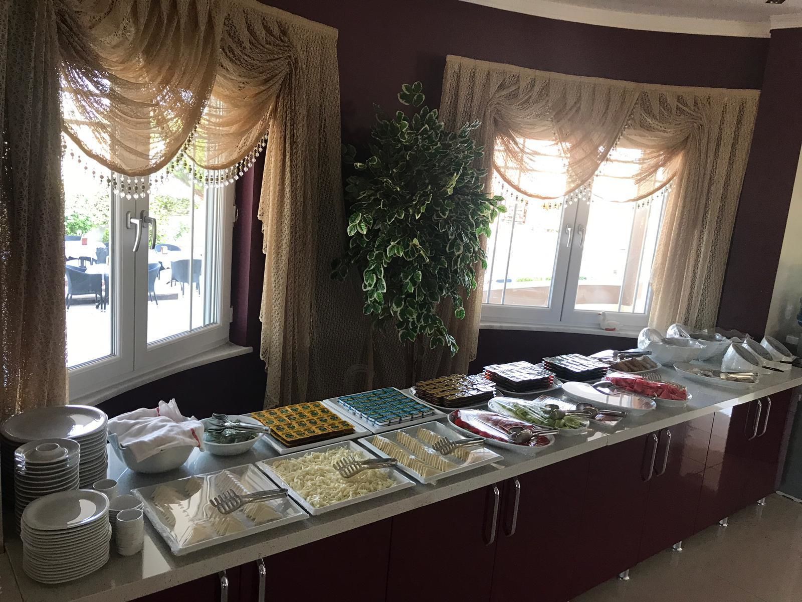 A buffet setup with various food items and utensils, a potted plant, and a window with curtains.