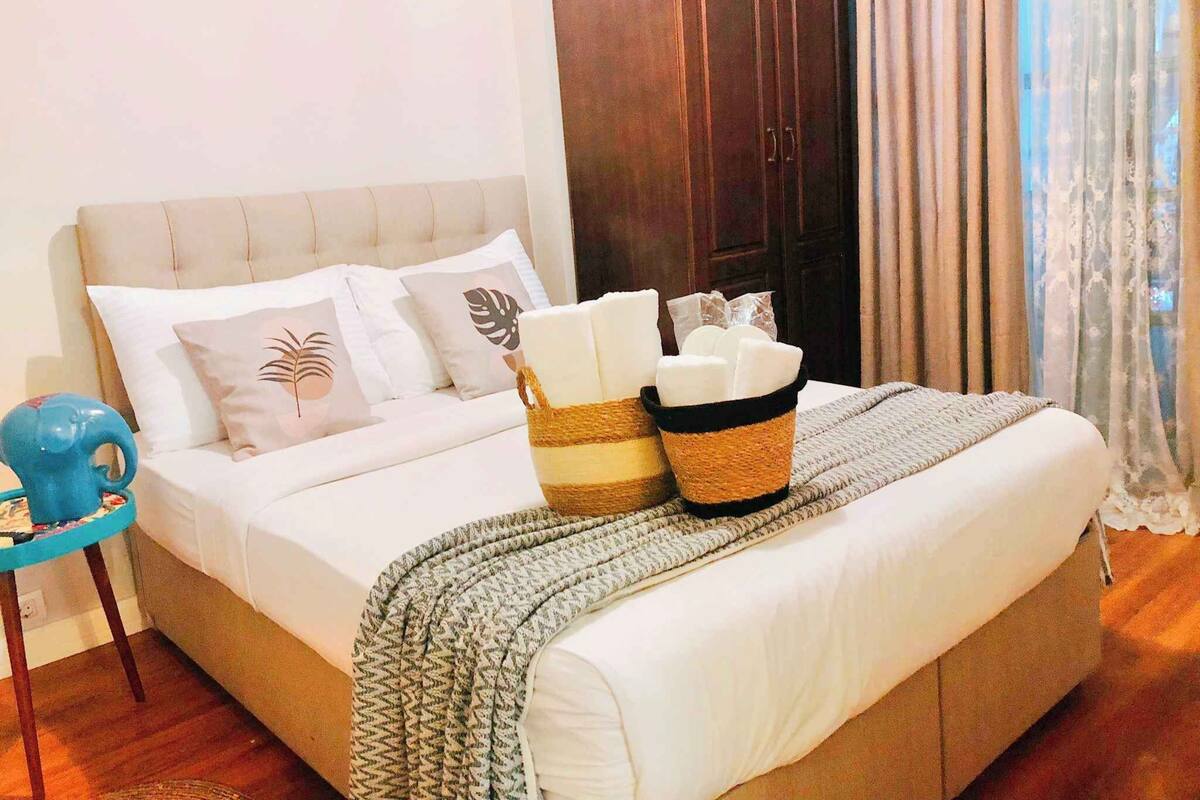 Een net opgemaakt bed met een beige hoofdbord, wit beddengoed en decoratieve kussens. Op het bed staat een rieten mand met kaarsen.