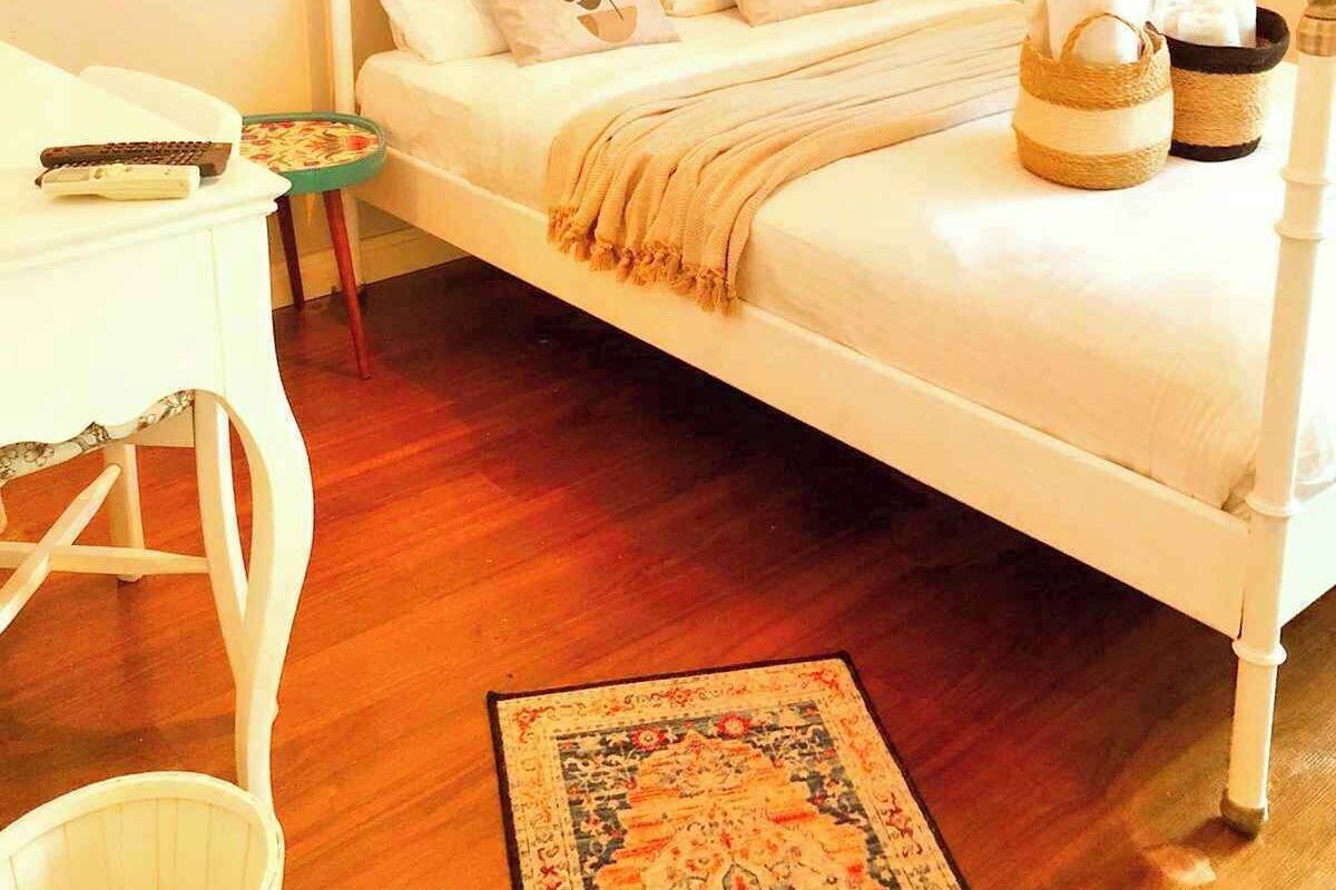 Een slaapkamer met een wit hemelbed, een klein nachtkastje, een stoel, een geweven mand met handdoeken en een macramé wanddecoratie.