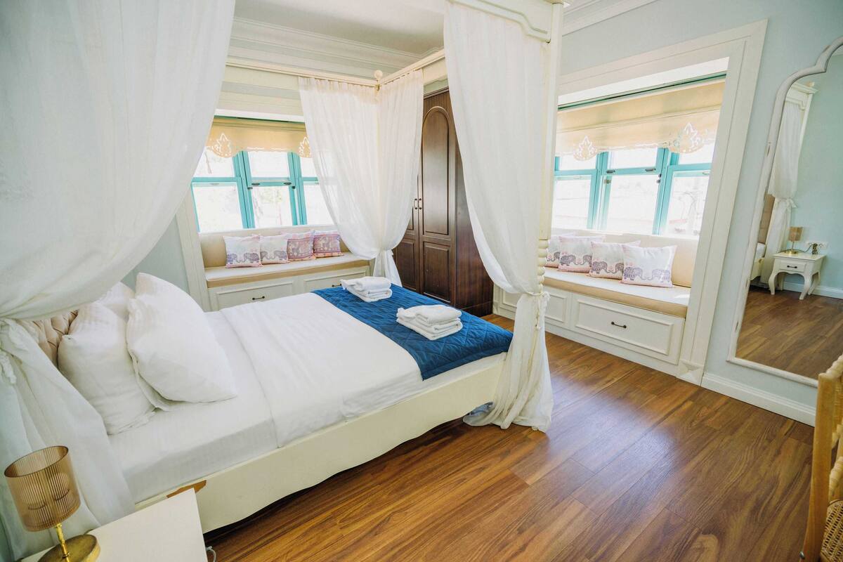 Een slaapkamer met een hemelbed, een vensterbank met kussens, een houten vloer en een spiegel.