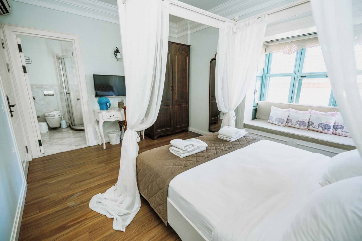 Een slaapkamer met een bed, een televisie, een kledingkast, een raam met gordijnen, en een badkamer zichtbaar door een geopende deur.