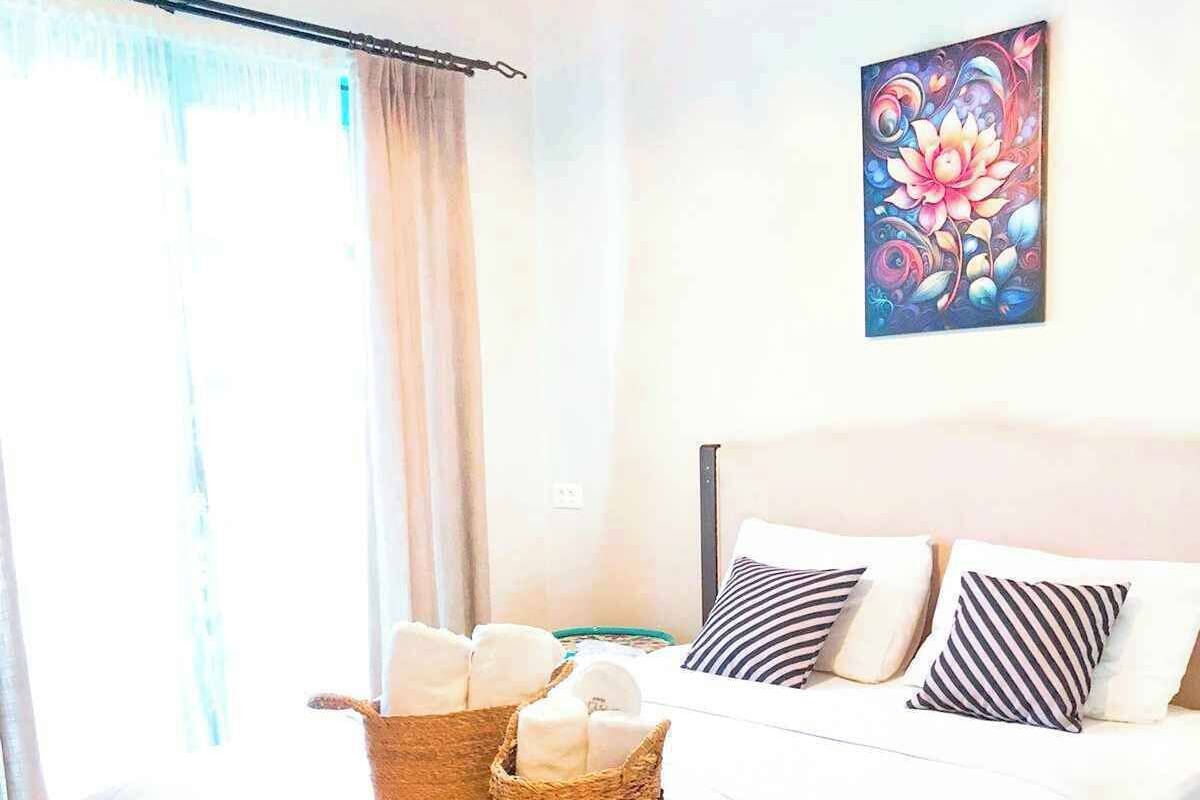 Een netjes opgemaakt bed met gestreepte kussens, een geweven mand met handdoeken en een kleurrijk schilderij aan de muur.