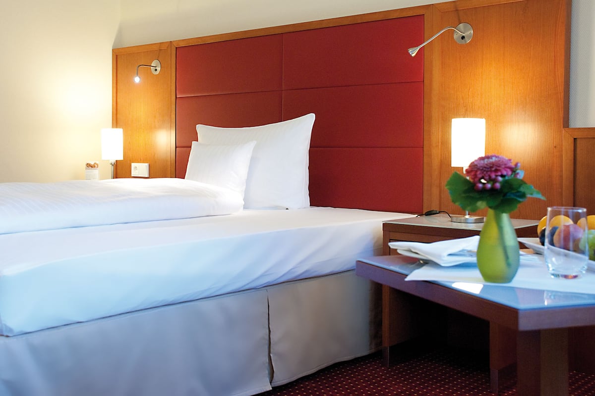 A hotel room with a large bed, a wooden headboard, two bedside lamps, a nightstand with a vase and a glass of water, and a red carpet.
