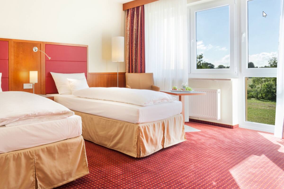 A hotel room with two beds, a desk, a chair, a window with a view of greenery, and a red carpet.