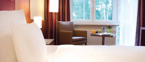 A hotel room with a large bed, a chair, a nightstand with a lamp, and a window with a view of trees.