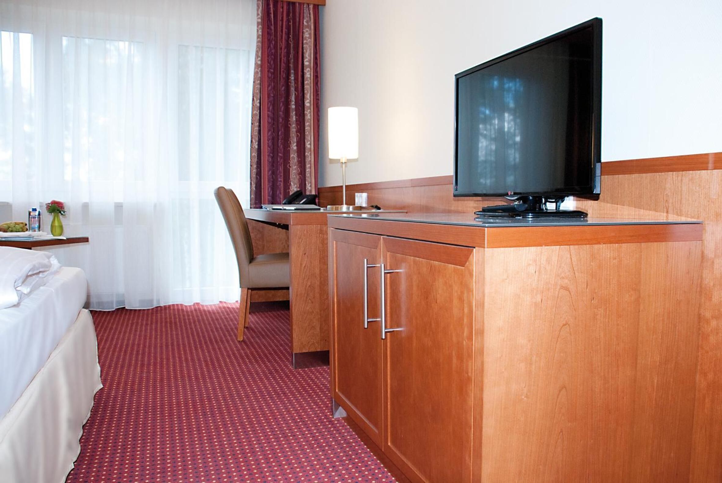 A hotel room with a bed, a desk with a television, a chair, and a small table with a vase and a bottle.