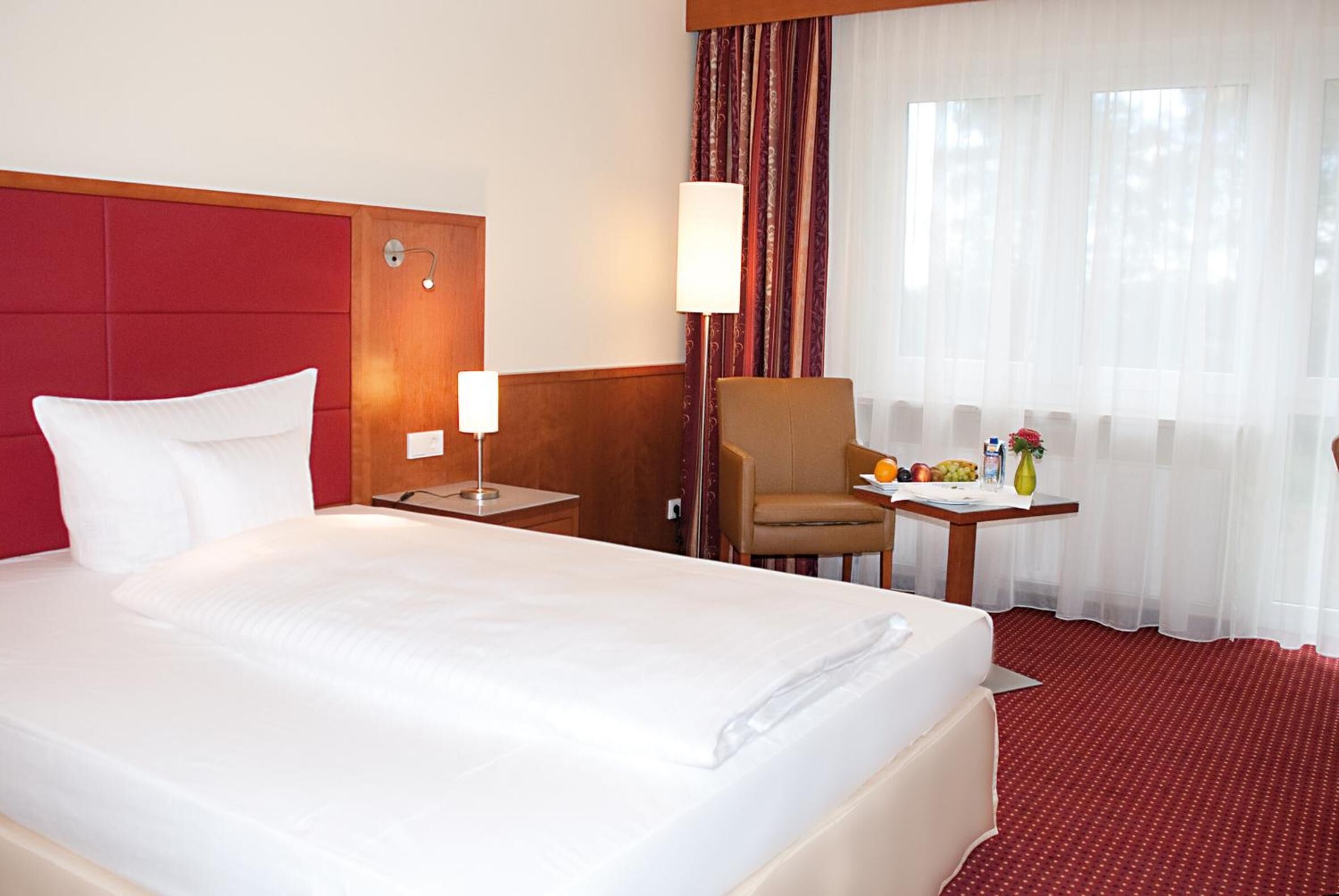 Hotelkamer met een groot bed, een houten hoofdeinde, een nachtkastje met een lamp, een stoel, een tafeltje met fruit en een vaas, en een raam met dunne gordijnen.