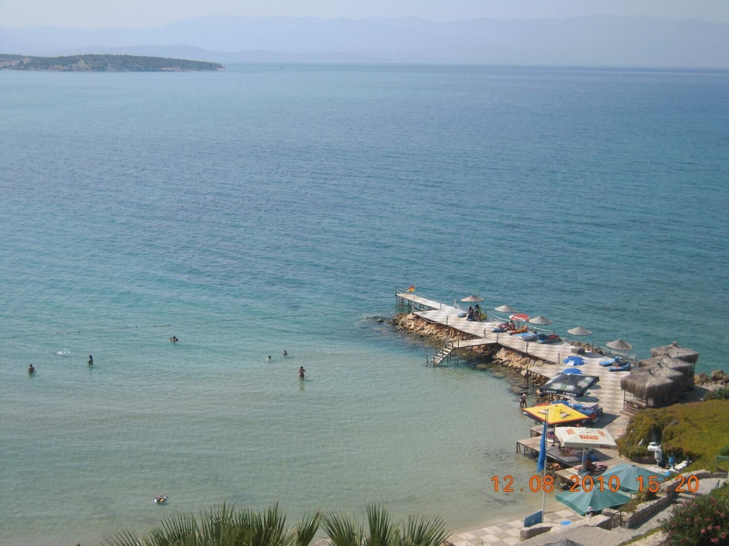 A coastal area with a pier extending into the sea, a beach with umbrellas, and people swimming.