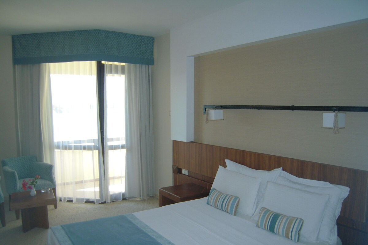 Quarto de hotel com cama, mesa de cabeceira, cadeira e uma janela grande com cortinas.