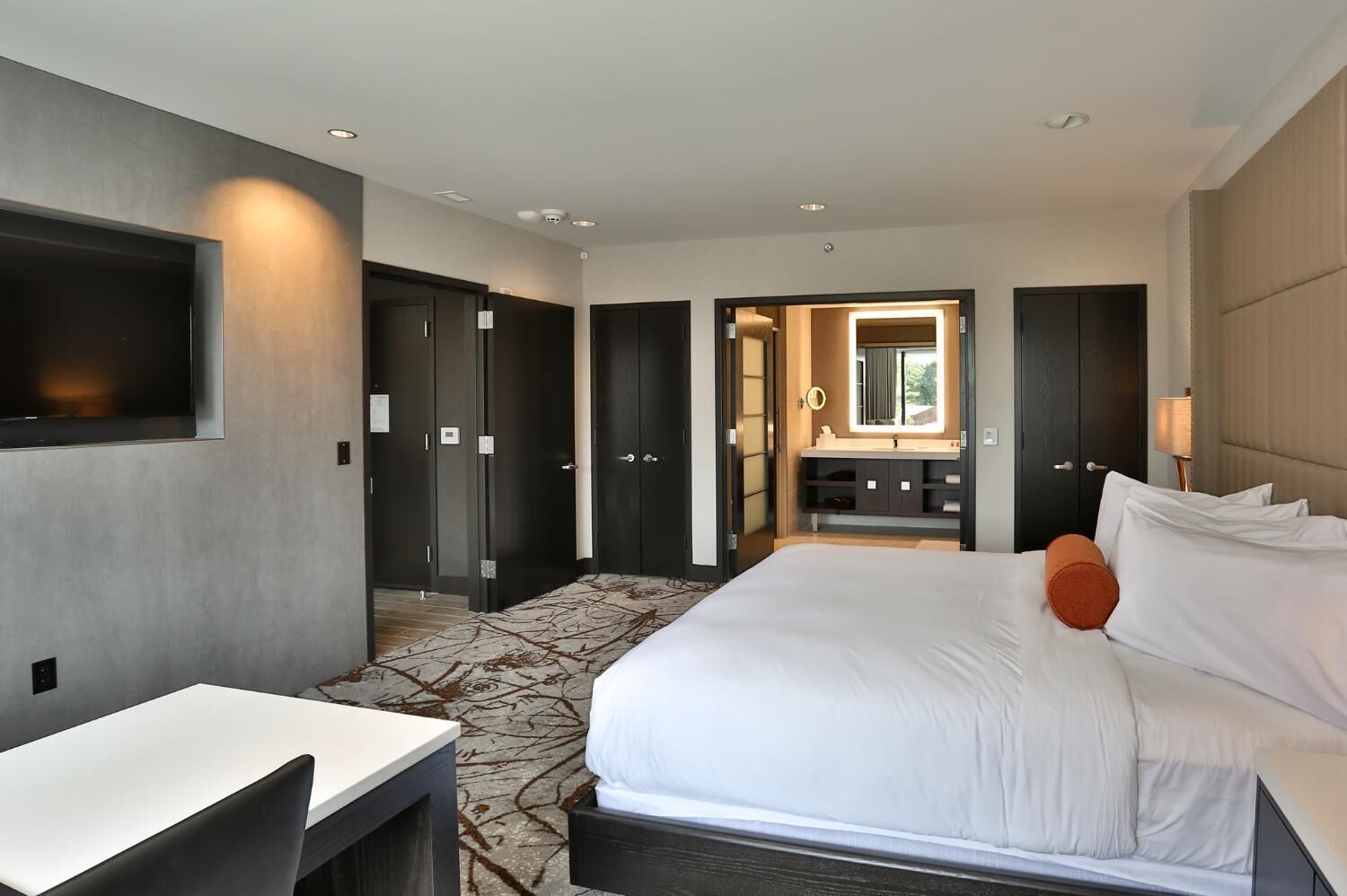 A hotel room with a bed, a desk, a mirror, and a bathroom visible through an open door.